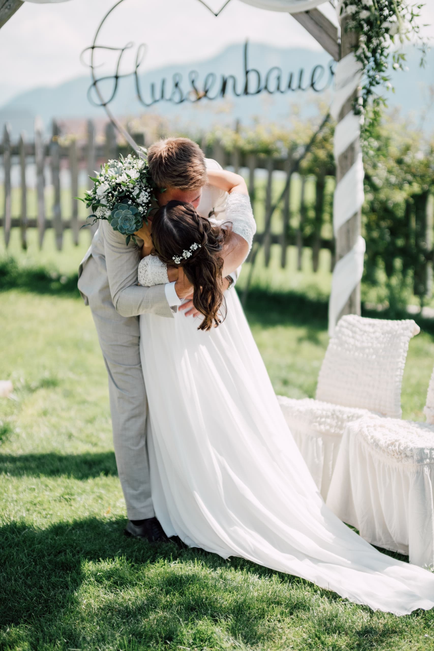 Groom embraces the bride under a wooden heart arch hung with white ribbons