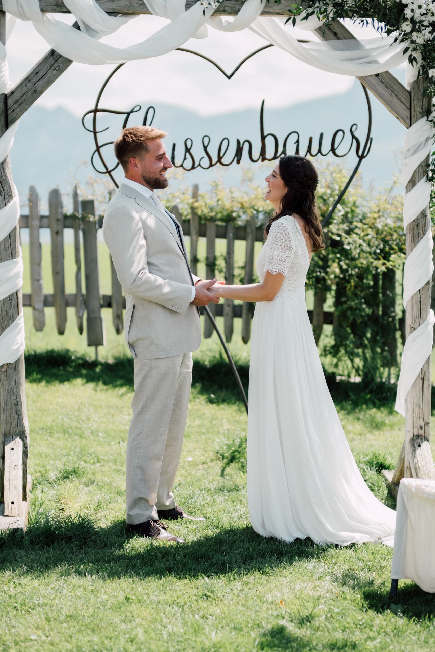 Groom and bride hold both hands facing each other under a decorated wooden ceremony arch