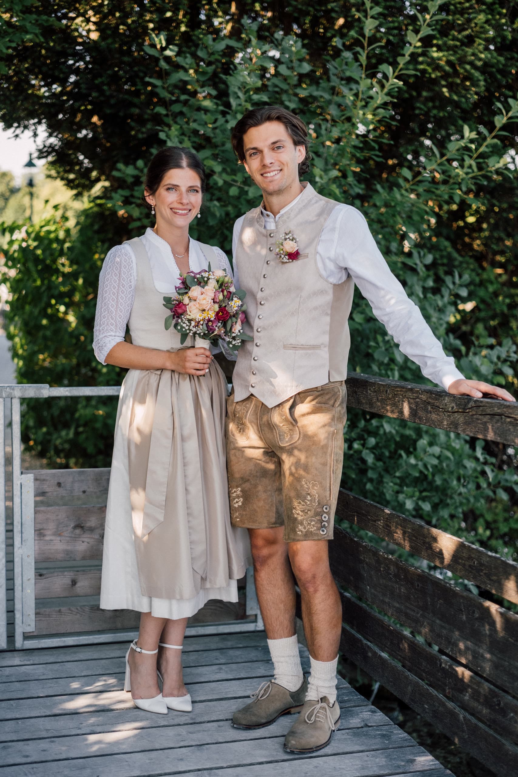 Couple in Austrian Tracht stand on a wooden deck before green foliage, she holds a bouquet