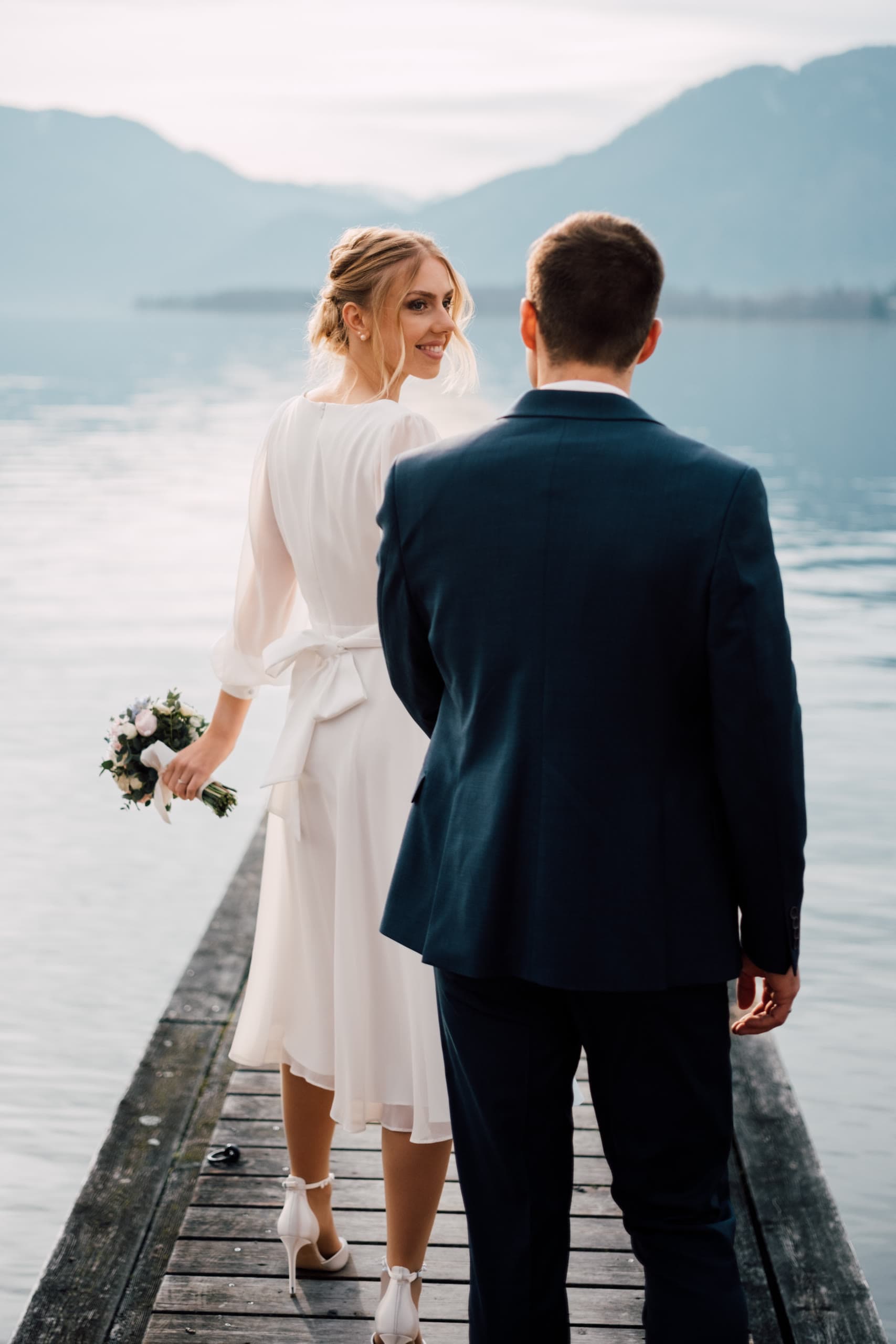 Bride looks back over her shoulder beside the groom on a lake dock at dusk
