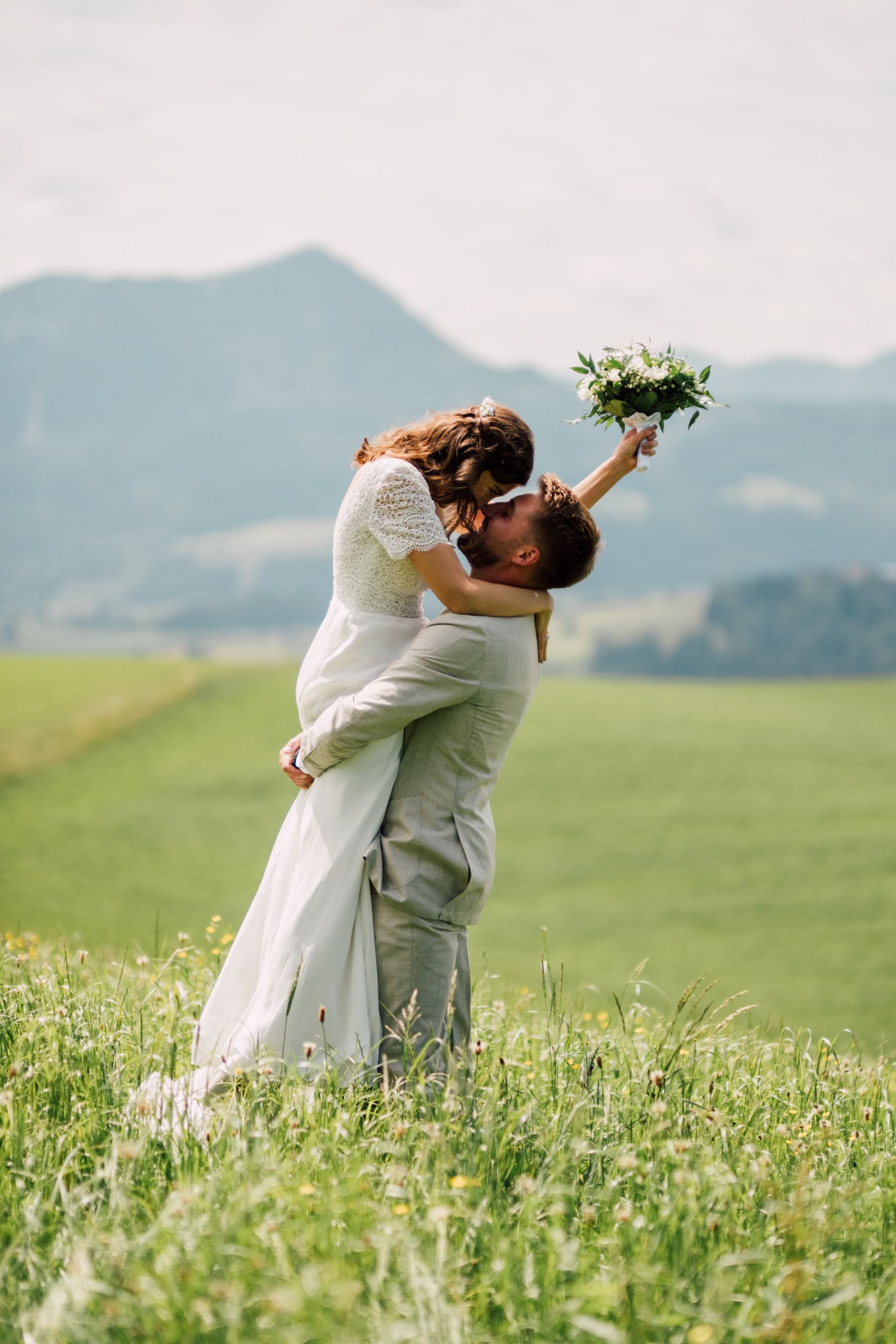 Groom lifts the bride as they kiss in a green meadow with mountains hazy in the distance