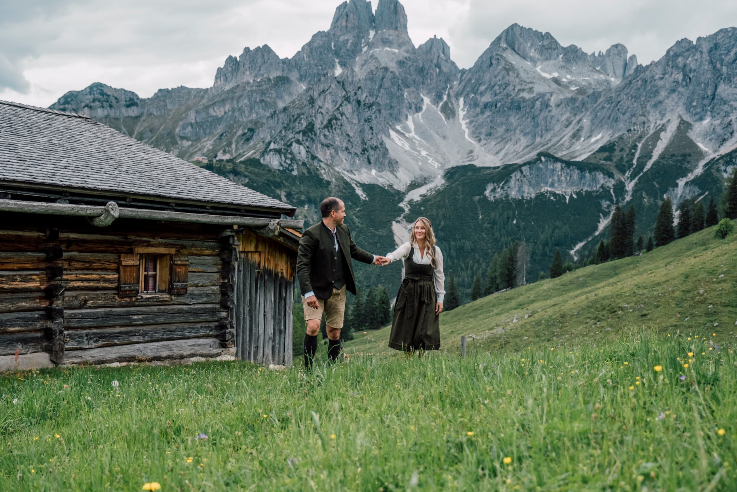 Paar in Tracht geht Hand in Hand über eine Wiese vor einer Holzhütte und grauen Gipfeln