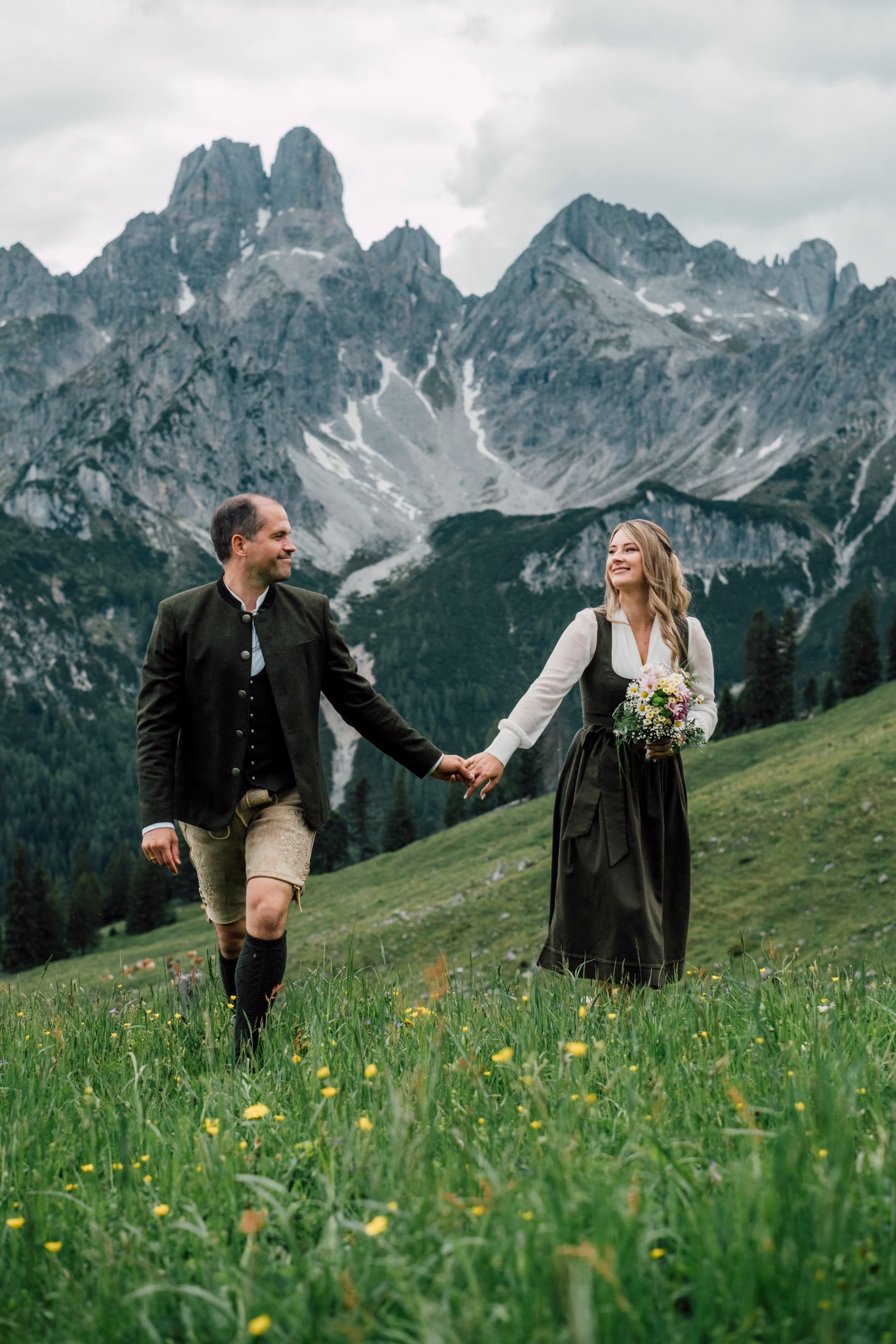 Couple in Tracht walk hand in hand through a flowering pasture below grey mountain peaks