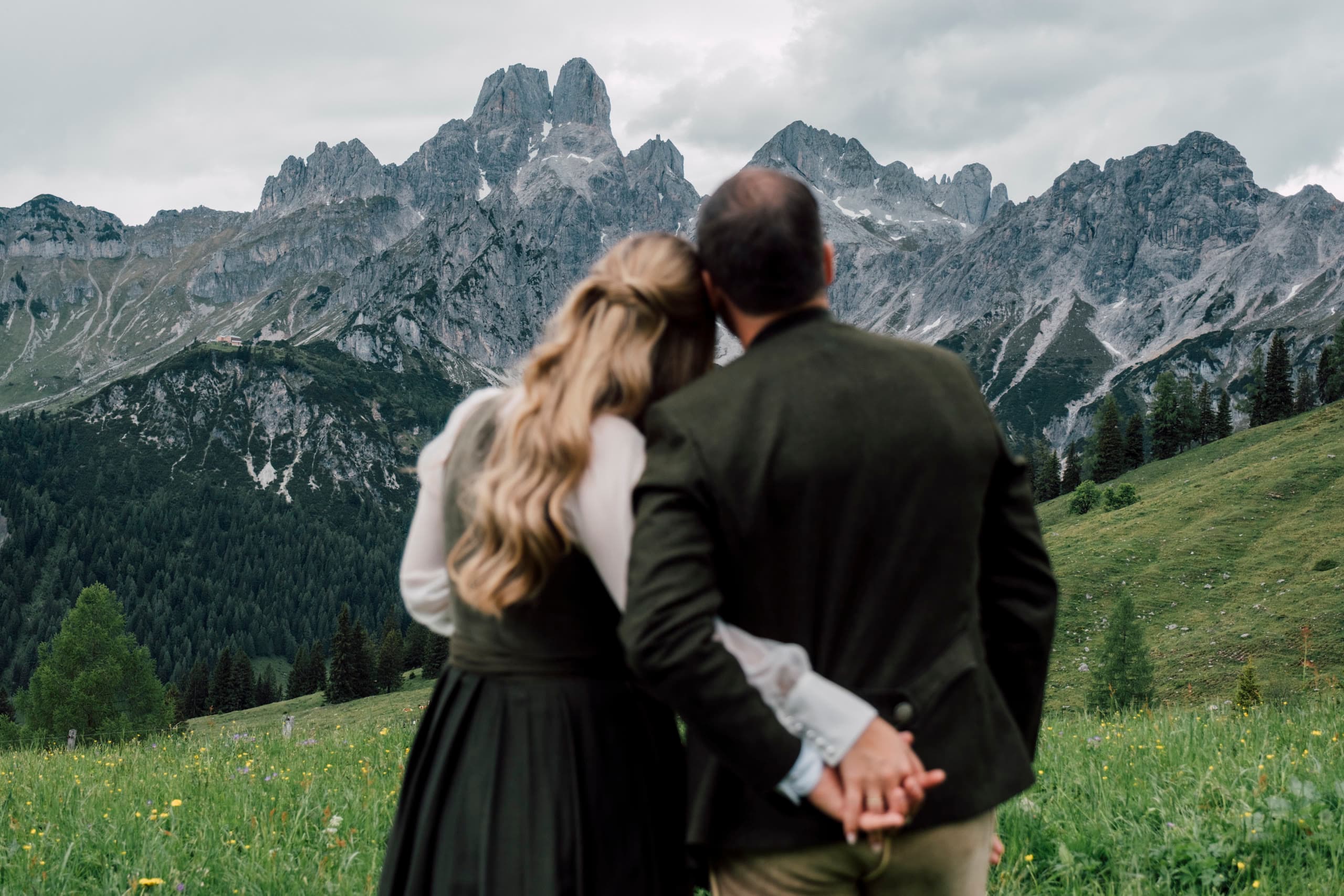 Couple stand hand in hand with their backs to the camera, facing grey rocky peaks