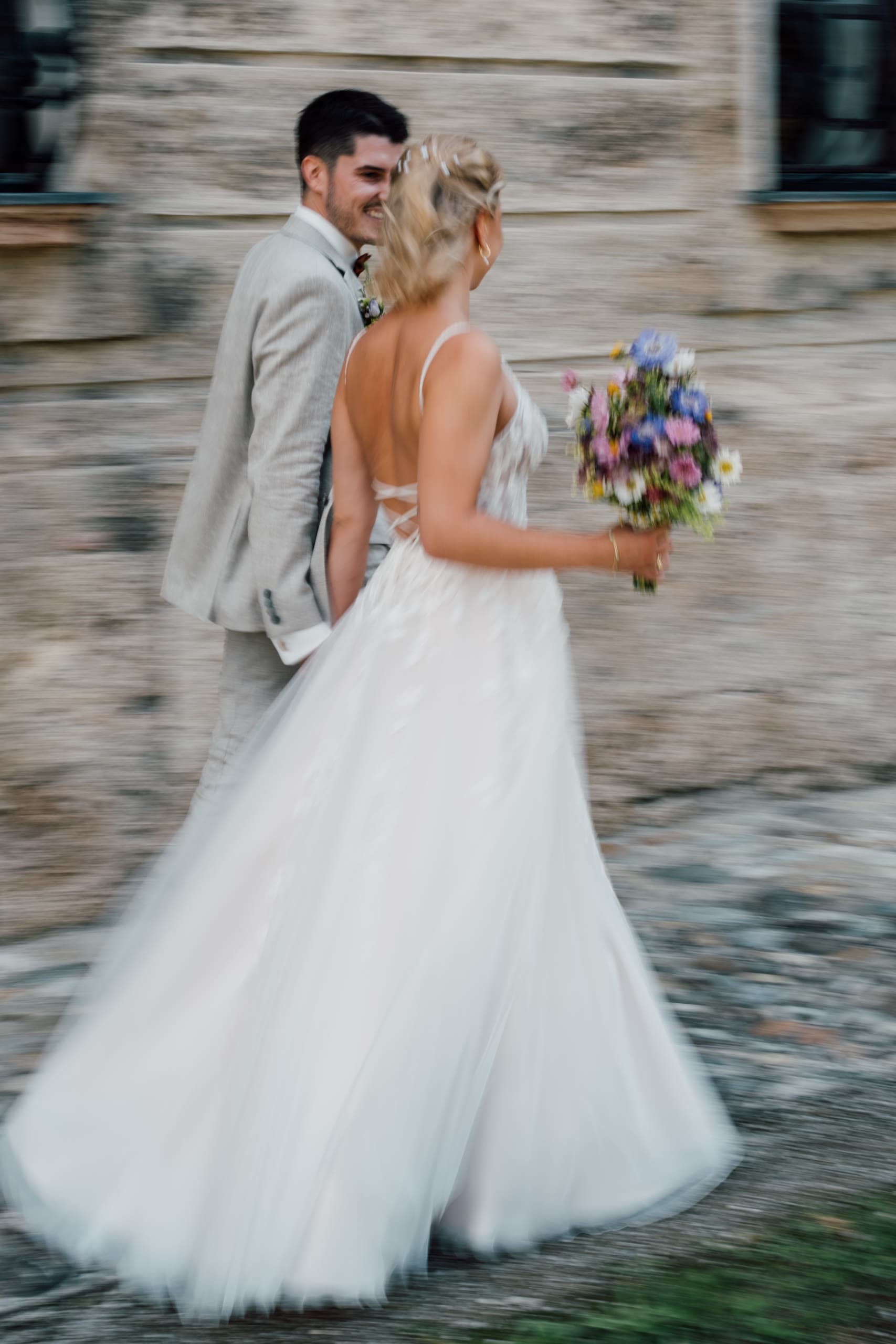 Bride glances back laughing while walking with the groom past a stone building