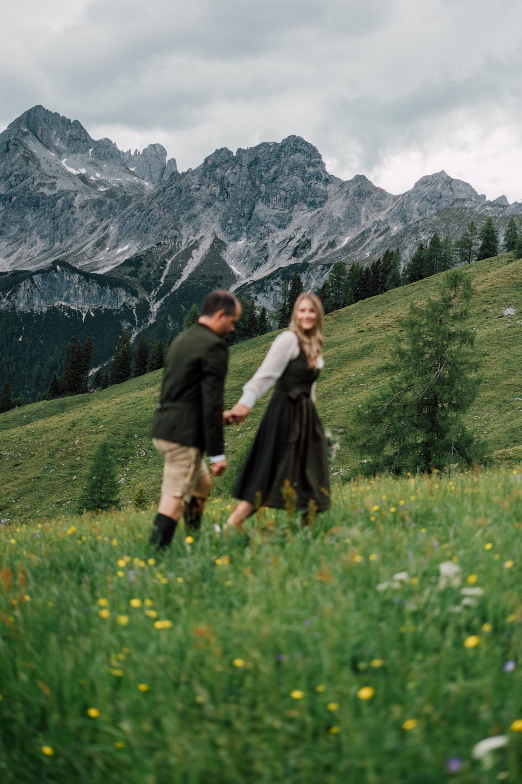 Couple in Austrian Tracht walk hand in hand through a wildflower meadow below mountains