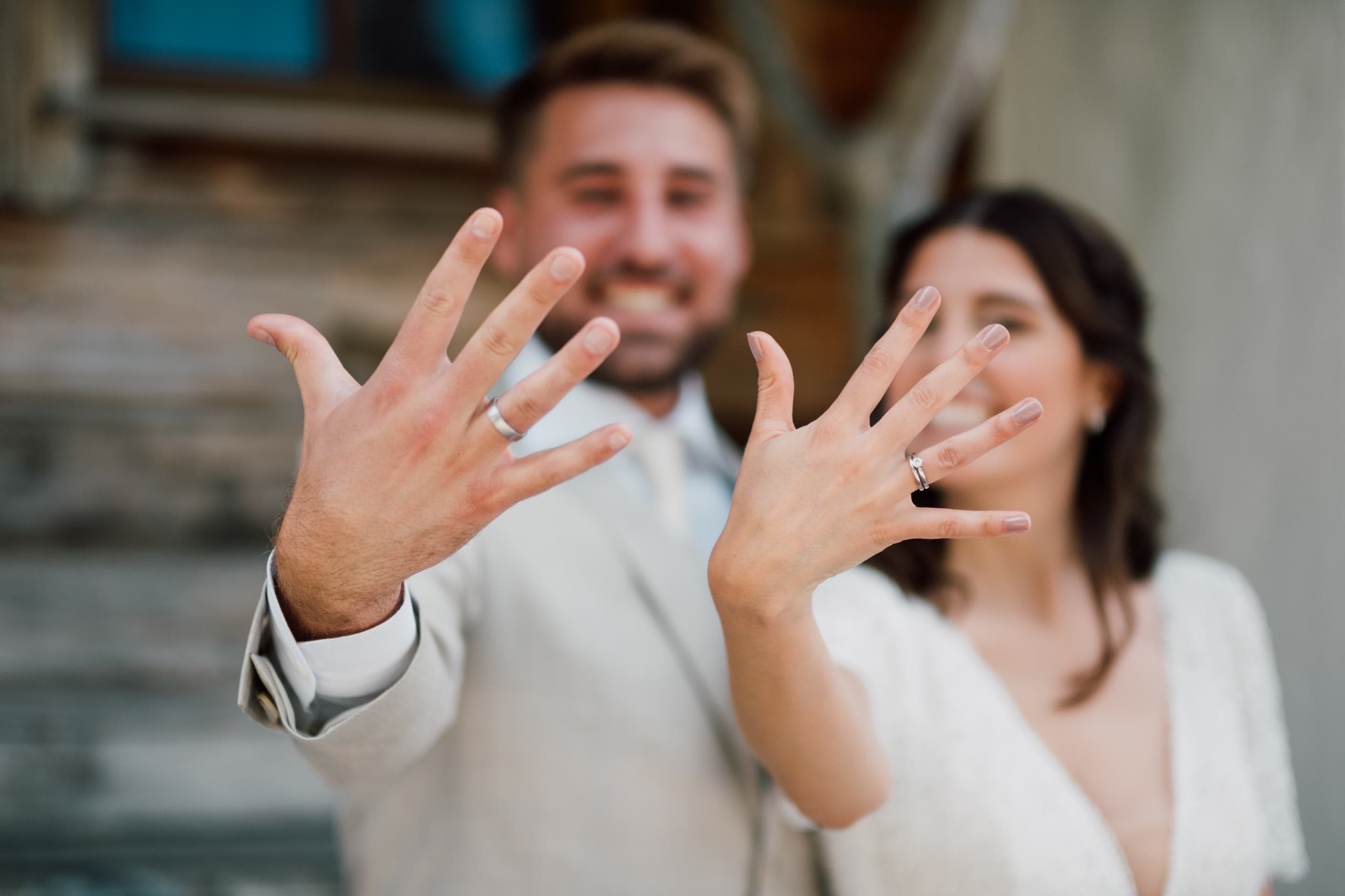Couple hold their hands up to the camera showing wedding rings, their faces blurred behind