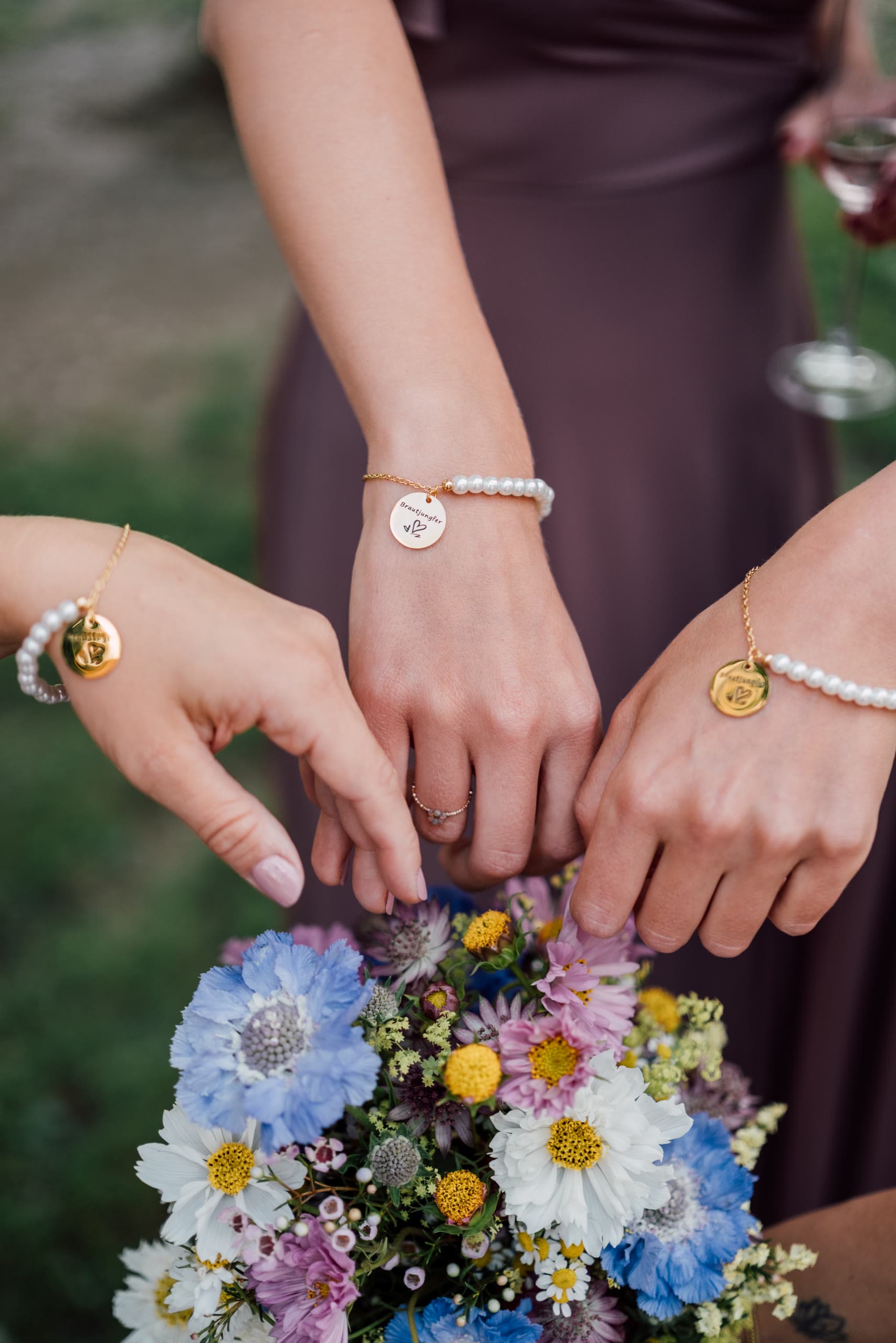 Three hands with pearl and gold bracelets reach to touch a bouquet of mixed wildflowers