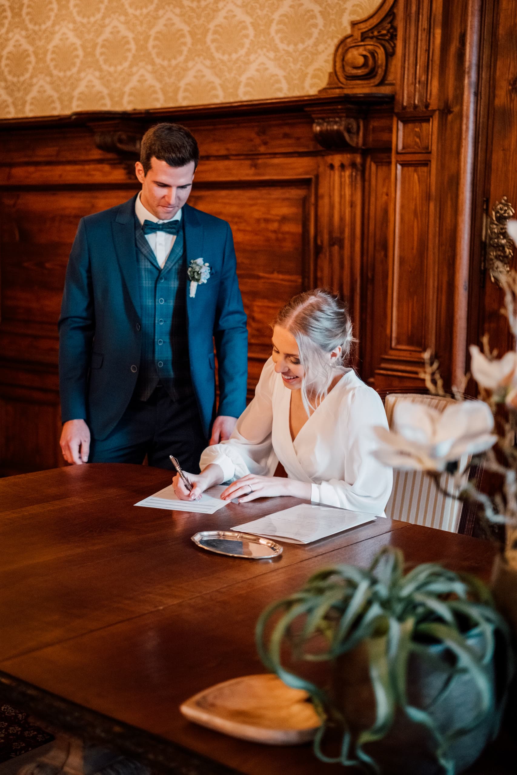 Bride signs a document at a wooden table while the groom in a teal suit stands beside her